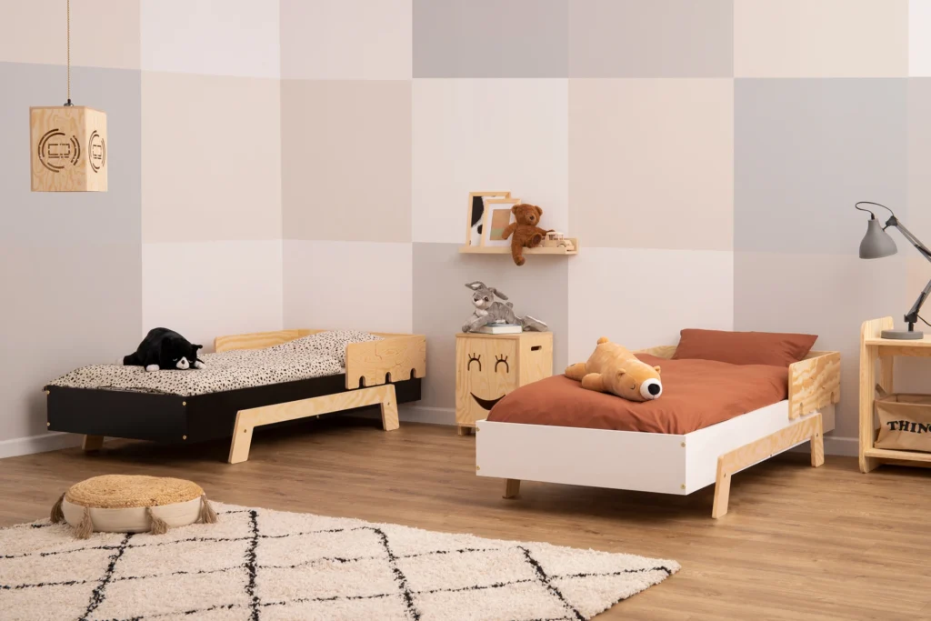 Deux lits en contreplaqué de pin avec des barrières en forme de puzzle dans une chambre lumineuse avec des peluches